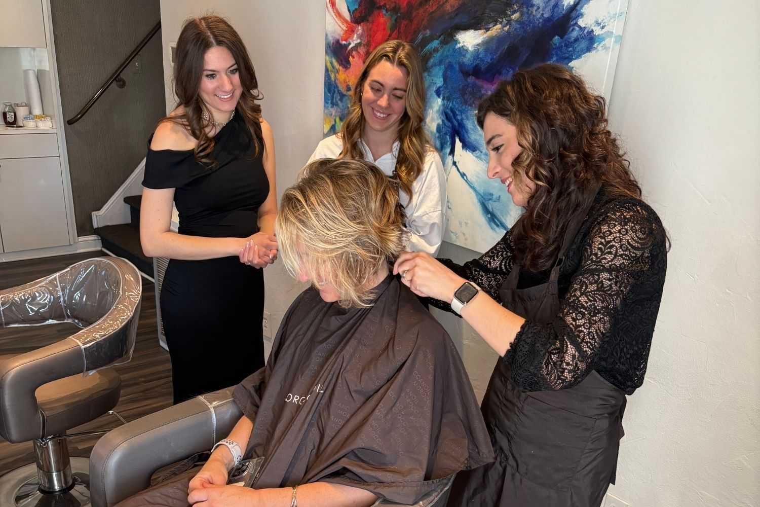 Hairdresser styling a smiling client's hair in a salon, watched by two others nearby.